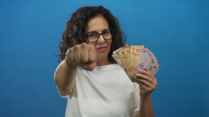 Middle age hispanic woman holds mexican pesos banknotes and extends fist to camera in blue studio; defiance empowerment.