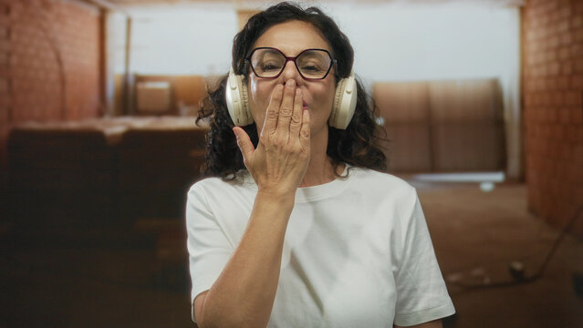 Woman wearing headphones blowing kiss in indoor construction site setting, exhibiting joy at work while interacting warmly with her environment.