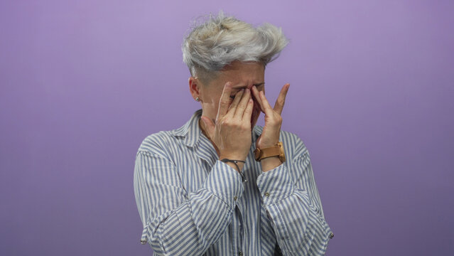Woman with short hair covers eyes with hands in purple studio set; sadness isolation grief despair.