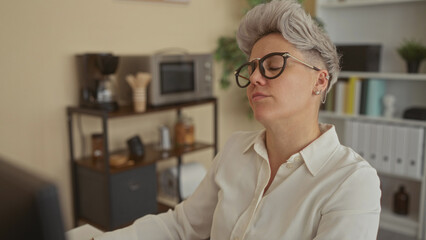 Woman with glasses leans forward at computer monitor on desk in office surrounded by shelves and a coffee machine; concentration.