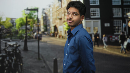 Fototapeta premium Young hispanic man wearing glasses and a blue shirt looks to the side showing his face on a city street; contemplation.