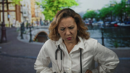 Woman doctor with stethoscope clutching stomach on street by an amsterdam canal, holding side in visible pain; medical concern.