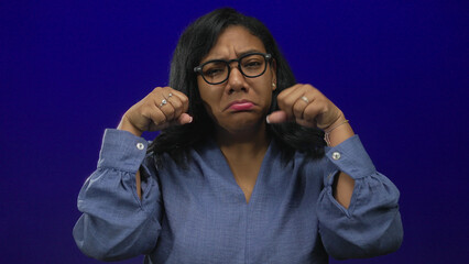 Obraz premium African american woman wearing glasses with frowning face and clenching fists in bright studio setting; sadness.