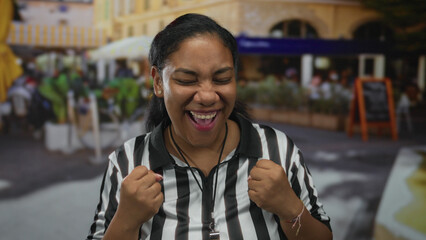Woman referee clenches fist in busy outdoor street soccer game in front of cafe; celebration joy...