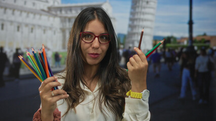 Young hispanic woman smiling outdoors at pisa tower holding colorful pencils in both hands against the iconic leaning structure with tourists in the background.