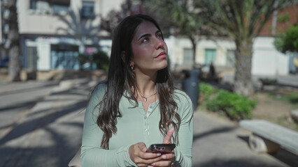 Fototapeta premium Young hispanic woman in green shirt using smartphone outdoors on a sunny urban street showing a serene and focused expression amidst a vibrant environment