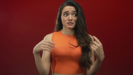 Woman shrugging with raised hands in orange crop top against red studio background; uncertainty doubt.