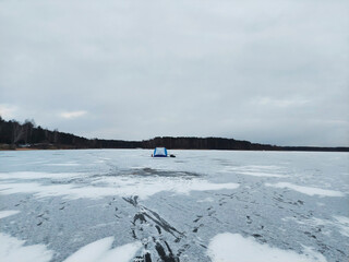 Ice fishing shelter on frozen lake during cloudy day in winter season with snow and distant trees on shoreline