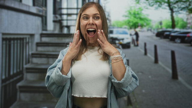 Woman wearing denim jacket over white shirt raises arms over her head on a busy street with open mouth; excitement.