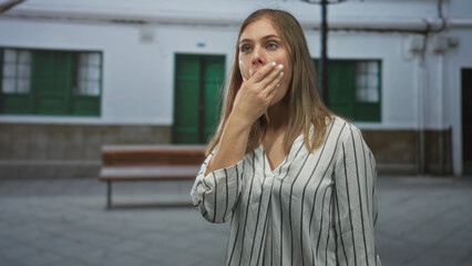 Woman holding hand to mouth with open mouth on street in front of white building and wooden bench;...