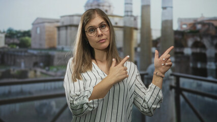 Fototapeta premium Blonde woman finger pointing to building facade under cloudy daylight on outdoor balcony railing by historic ruins; curiosity.
