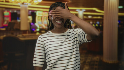 Woman covering eyes with hand and smiling at casino slot machines on indoor gaming floor; playful surprise.