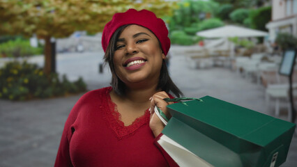 Woman smiling in red clothing holds shopping bags on a city terrace with outdoor seating and greenery, showcasing youthful joy and urban shopping experience.
