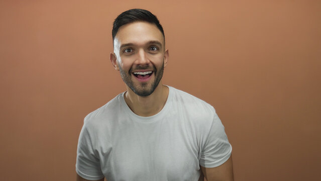 Hispanic young man in casual white shirt with short hair smiling against orange background conveying positivity and energy