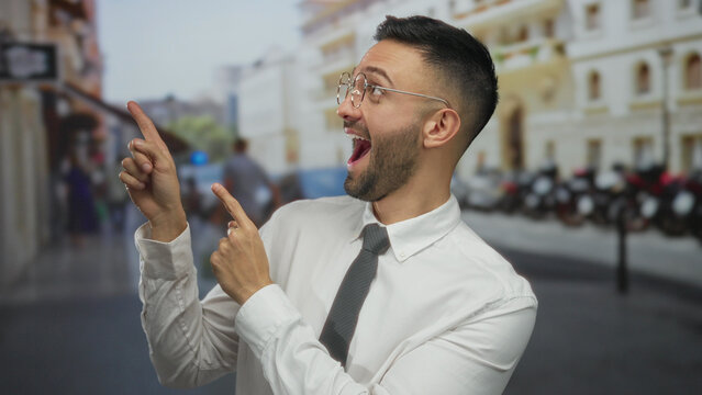 Hispanic man in white shirt and tie expressively pointing in an urban city street setting with blurred buildings and people in the background, wearing glasses and appearing surprised.