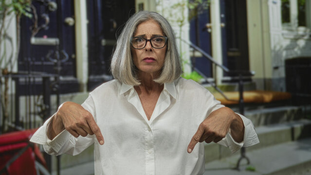 Woman points fingers downward at her shirt on building steps with iron railing and stoop; frustration.