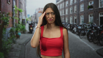 Fototapeta premium Young hispanic woman in red crop top holding hand to temple and frowning with headache while standing on a narrow city street lined with parked bicycles; discomfort.