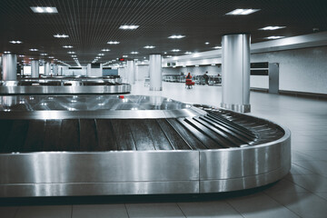 Modern airport baggage claim area with stainless steel luggage carousel, tiled floor, bright ceiling lights and columns, empty terminal hall for travel arrivals
