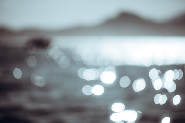 Abstract defocused seascape with sparkling bokeh highlights on water, blurred mountain silhouette and pale blue sky, soft selective focus background with copy space