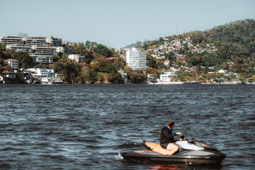 Wide view across bay showing coastal city on green hillside with modern apartment blocks and homes, blue sky and choppy water, with a silhouette of a jet ski rider speeding in defocused foreground