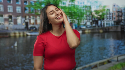 Fototapeta premium Woman in red shirt hand on cheek smiling on street by a canal with brick buildings in amsterdam; joy travel moment.