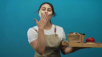 Woman waitress in apron holding a wooden tray with takeaway boxes and an apple, blowing a kiss with hand to lips in studio; warm hospitality cheerful.