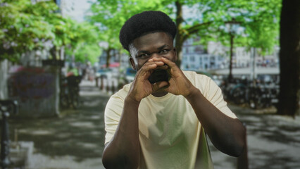 Young african american man cupping hands to mouth shouting while seated at a cafe table on a leafy...