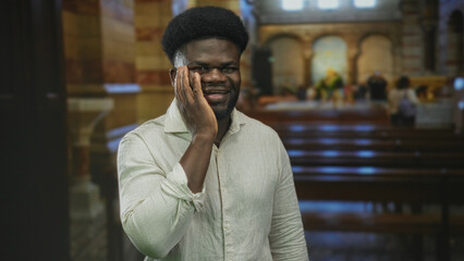 Obraz premium Man with hand on cheek smiling and looking ahead inside a church building beside wooden pews, wearing light linen shirt and standing near altar; faith reflection joy.