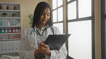 Woman doctor in a clinic room reviews a clipboard, surrounded by medical supplies and daylight streaming through large windows.