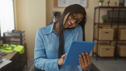 Woman multitasking in an office holding a tablet, wearing glasses, and smiling while using a phone and pen, suggesting a busy work environment.