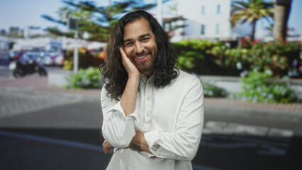 Fototapeta premium Man with long hair and beard smiling, hand to cheek and folded arm on city street near planter and parked scooter; shy joy.