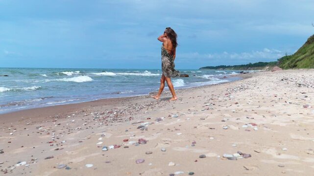 woman in flowing dress walking along shellstrewn beach, breeze tousling hair, ocean waves and pebbles underfoot, contemplative