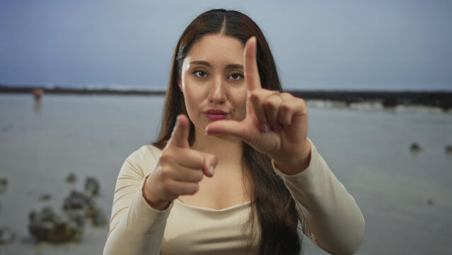 Woman holds a loser sign with her hand toward the camera on a windy beach with scattered rocks under an overcast sky at low tide; mockery.