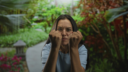 Woman clenching fists toward camera in forest park path near stone lantern and flowerbed, face tense with furrowed brow and hands raised; frustration.