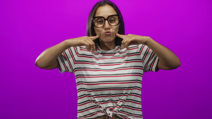 Woman presses cheeks with fingers while wearing striped shirt and oversized glasses in magenta studio; playful confident youth.
