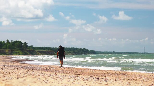 male traveler walking along sandy shoreline with backpack and surf, blue sky with scattered clouds, contemplative pace and distant