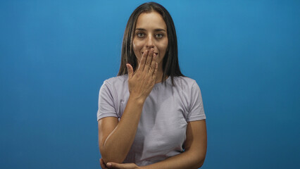 Young hispanic woman covering mouth with hand in blue studio, wearing light purple t shirt and...