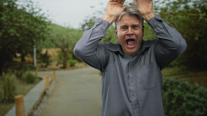 Man with grey hair holding hands on head in playful antler gesture on a tree lined paved street,...