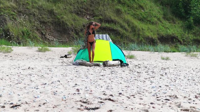 bikini woman walks across sandy beach beside colorful tent, green cliff backdrop, scattered pebbles, woman adjusts sunglasses