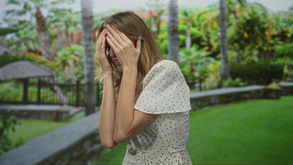 Woman covering face with hands in a forest park near a gazebo and stone wall, wearing a white polka dot dress; anxiety vulnerability.