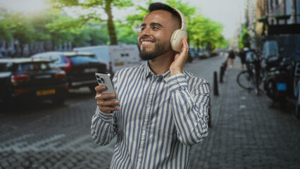 Young hispanic man smiling, holding smartphone in one hand and touching headphones while standing on a cobblestone street; music joy escape.