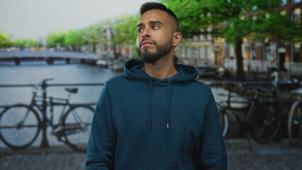 Fototapeta premium Man with hands on cheeks for toothache on amsterdam canal street near bicycles and bridge, wearing hoodie, looking pained; urban worry.