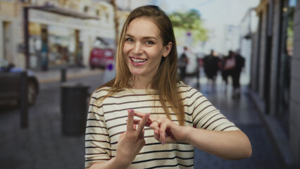 Woman forming hashtag sign with fingers on street while smiling brightly under sunlight and busy storefronts; playfulness.