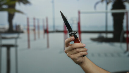 Hand holding knife outside gym with blurred palm trees in background, implying intense atmosphere of man in outdoor fitness setting.