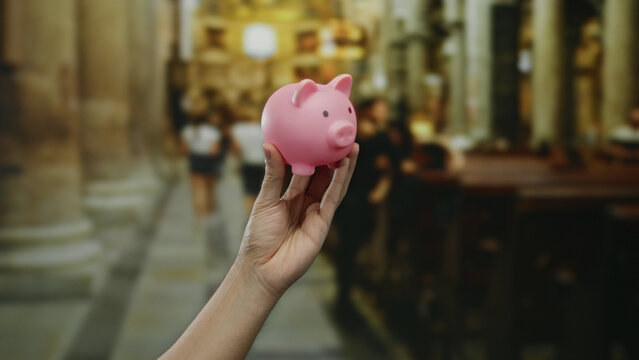 Man holding pink piggy bank in church, highlighting savings and finance in a religious setting with blurred congregation in background.