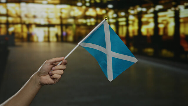 Hand holding scottish flag in city street showcasing national pride outdoors at night with blurred lights in the background.