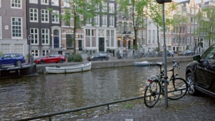Fototapeta premium Bikes parked along the picturesque amsterdam canal under a blurred, defocused background of traditional dutch houses and vibrant trees reflecting in the water.