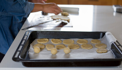Asadera o placa para horno con papel manteca donde reposan galletitas caseras sobre la mesada de la cocina, detras se ven las manos amasando