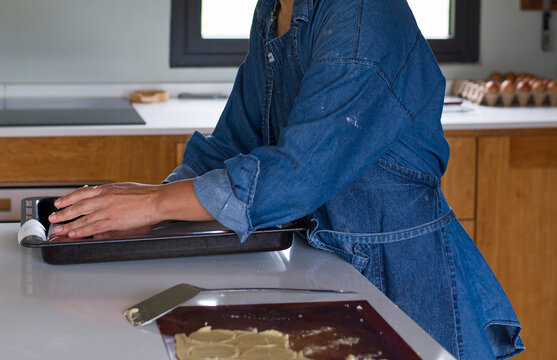 Mujer de piel morena con delantal azul jean pone papel manteca para hornaer galletitas sobre la mesada de la cocina