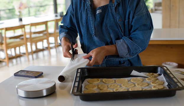 Mujer morena con delantal azul corta con tijeras papel manteca junto a ella hay una asadera o placa para horno con galletitas caseras sobre la mesada de la cocina